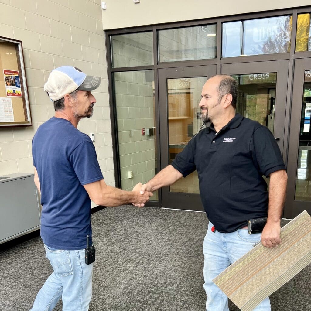 flooring expert holding a large vinyl plank shaking the hand of a client outside a building